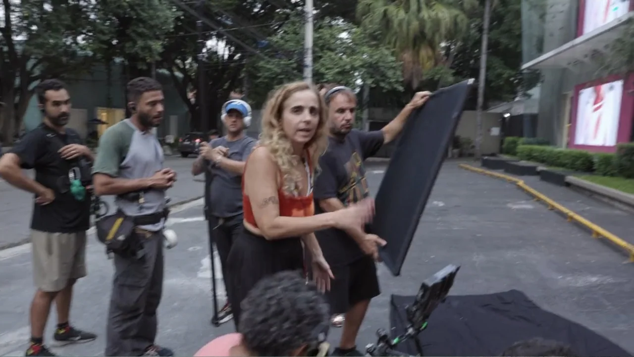 Making of: Scars of Beauty (2025) movie still: medium — A group of film crew members stands on an asphalt urban lot. A woman in an orange top holds…