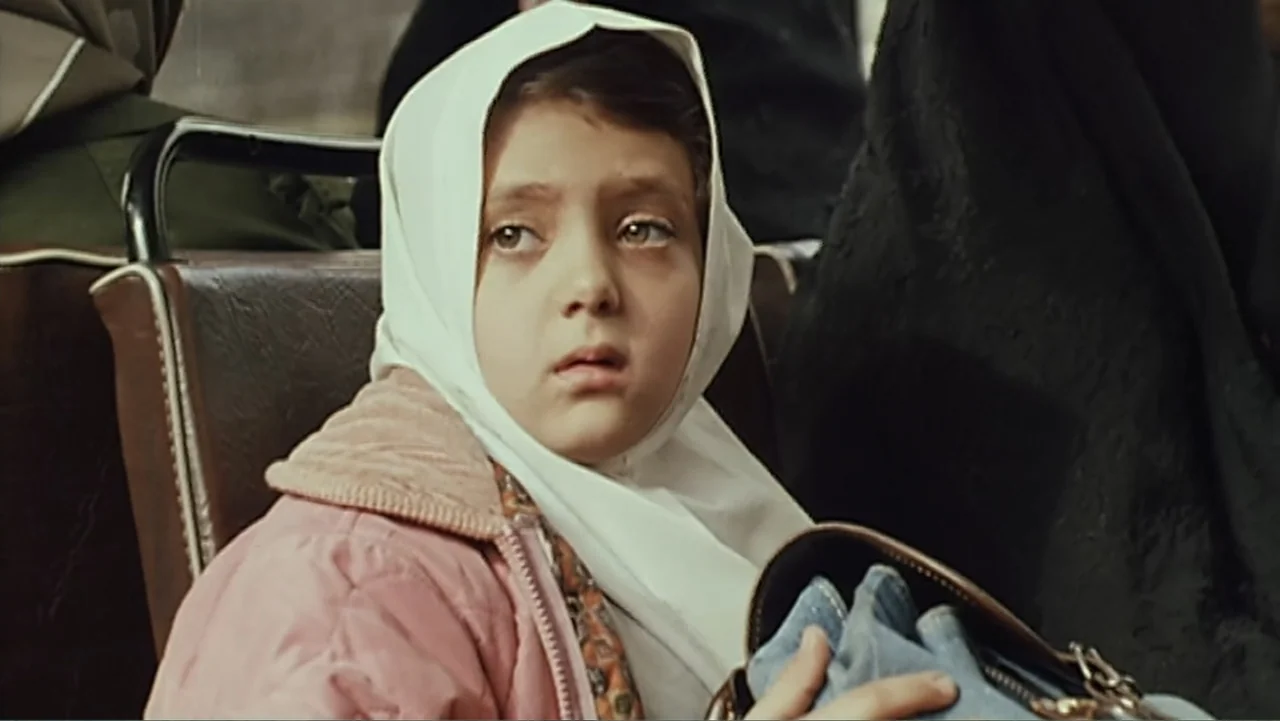 The Mirror (2014) movie still: medium — A child sits on a bus seat with a brown headrest and metal railing. The girl looks forward while holding a…