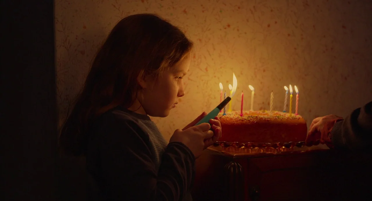 Petite Maman (2021) movie still: medium — A dimly lit room features a cake with burning candles on a wooden table. A young girl is in profile using…