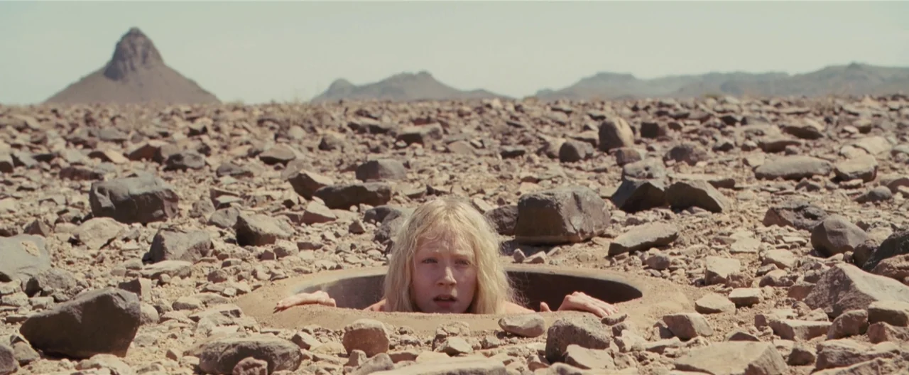 Hanna (2011) movie still: medium — A vast, rocky desert plain stretches out under a clear sky. A young woman emerges from a circular opening…