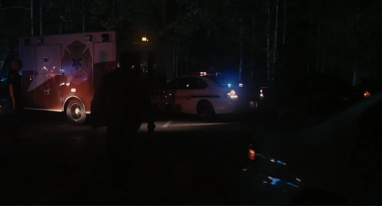 Beyond a Reasonable Doubt (2009) movie still: wide — A dark parking lot at night is bordered by dense forest trees. A person walks toward a parked…