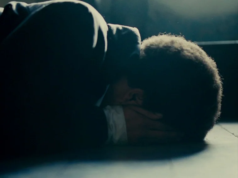 Wuthering Heights movie still: closeup — A dark indoor space where a person lies prone on a wooden floor. The individual rests their head on their…