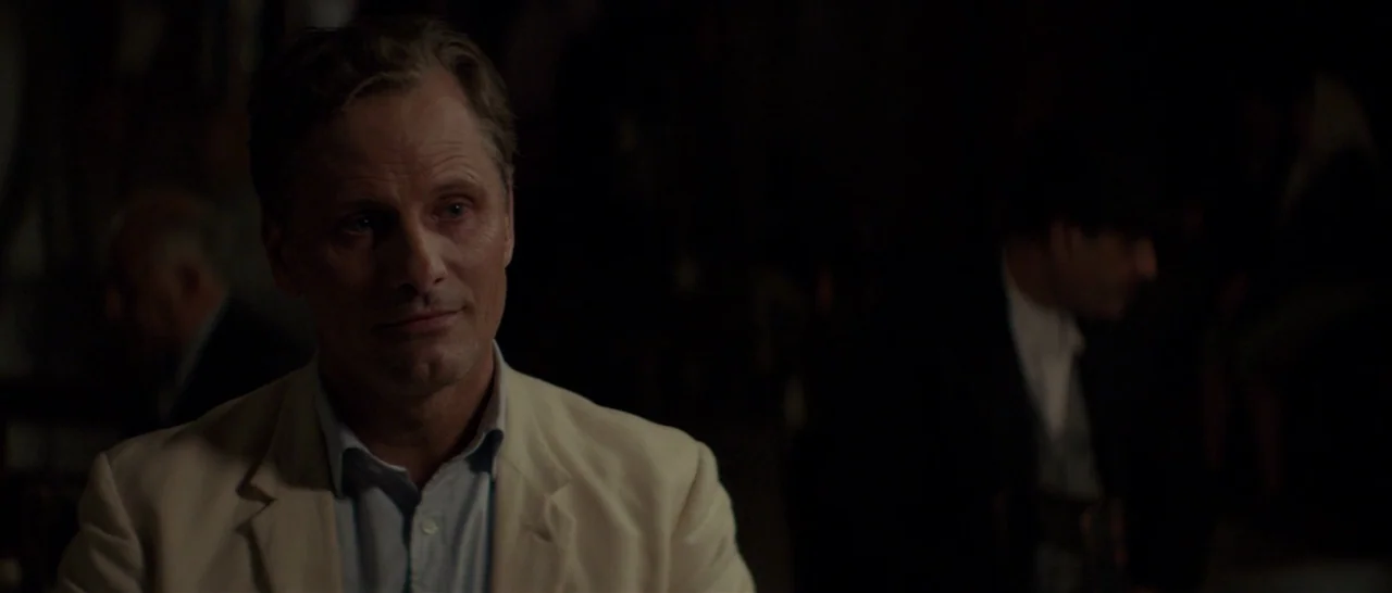 The Two Faces of January (2014) movie still: medium — A dimly lit room features a man sitting at a table. He wears a light suit while looking toward…