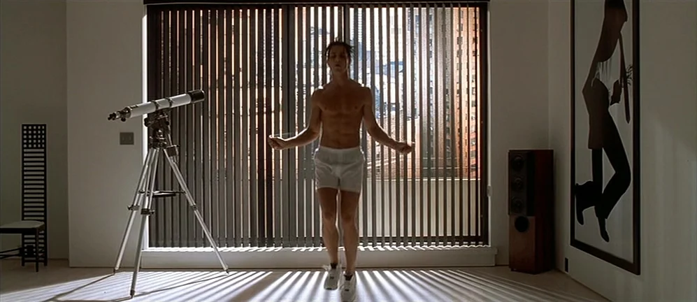 American Psycho (2000) movie still: fullbody — A minimalist modern apartment with floor-to-ceiling windows covered by vertical blinds. A man jumps…