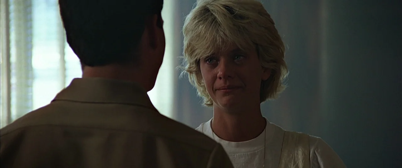 Top Gun (1986) movie still: medium — An interior space with a window partially covered by vertical blinds. A woman faces an unseen man standing with…