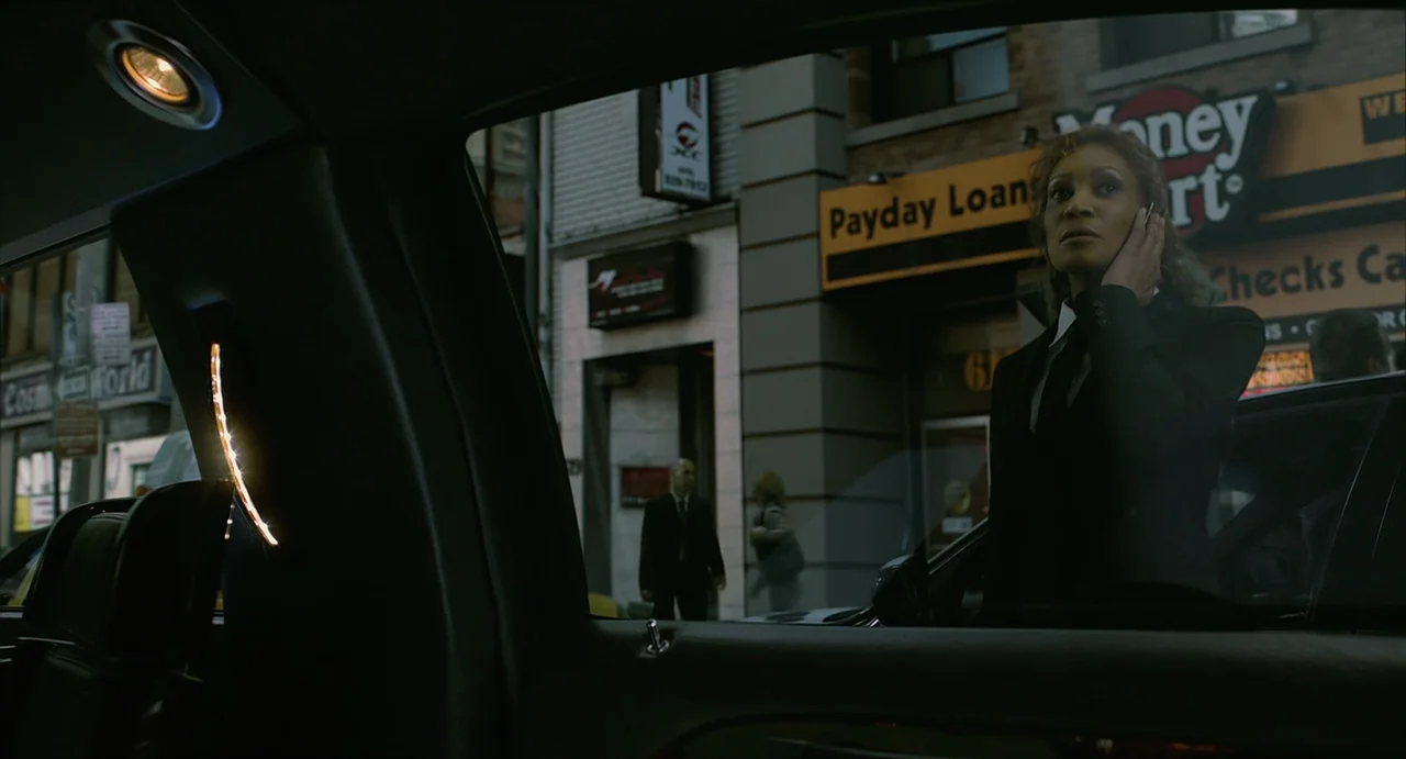 Cosmopolis (2012) movie still: medium — A view from inside a car looking out at a city sidewalk. A woman in a black suit talks on a phone outside…