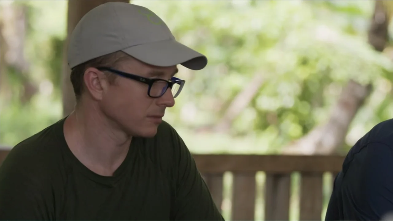 Finding Amelia (2024) movie still: medium — An outdoor wooden pavilion with blurred foliage visible in the background. A man wearing glasses and a…
