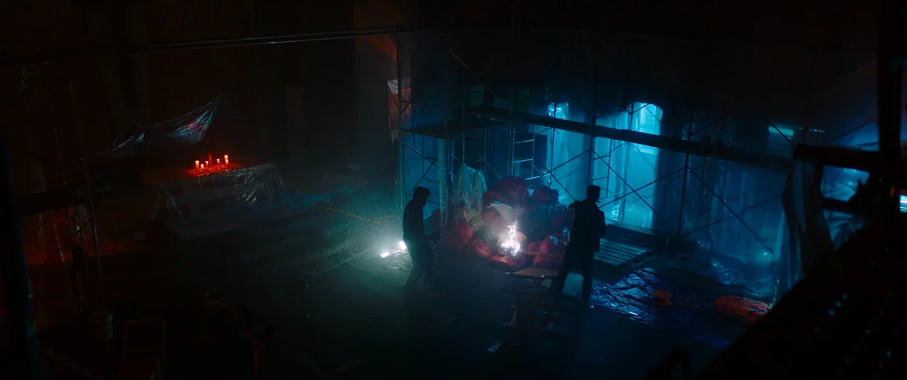 The Divine Fury (2019) movie still: wide — A dark church interior under renovation features metal scaffolding, plastic drop cloths, and red candles.…