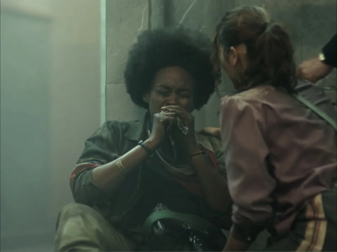 November (2025) movie still: medium — A dimly lit concrete stairwell is filled with light haze and architectural shadows. One woman sits on the…