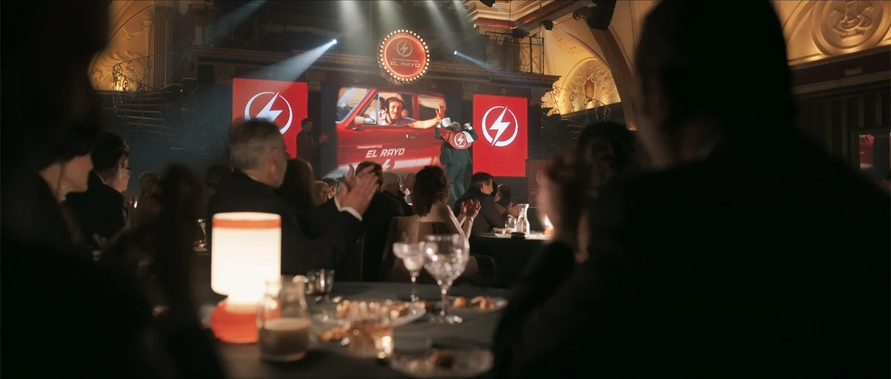 Historias lamentables (2021) movie still: wide — A formal event takes place in an ornate room with dark tables and patrons. A presenter stands on a…