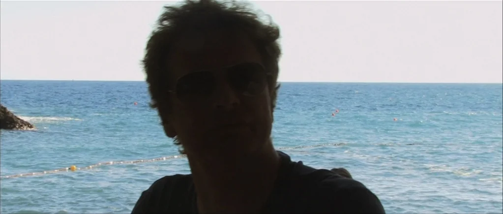 Genova (2008) movie still: medium — A man stands outdoors with the Mediterranean sea visible in the background. Bright daylight silhouettes the…