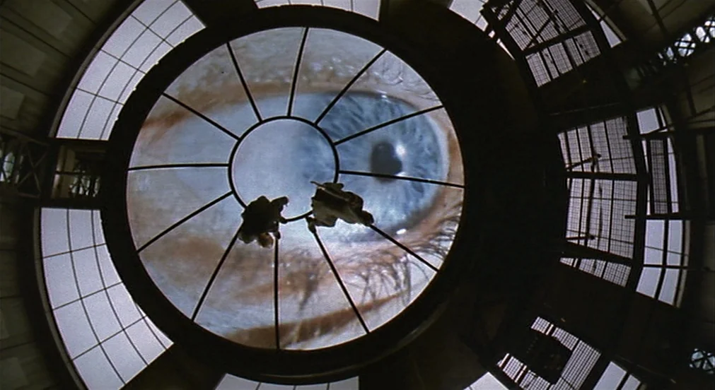 Kafka (1991) movie still: wide — A circular dome structure with metal framework overlooks a massive projected human eye. Two small figures appear as…