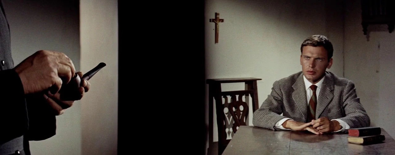 And God Created Woman (1956) movie still: medium — A man sits at a wooden table in a dimly lit room with a crucifix. Beside him, a person in dark…