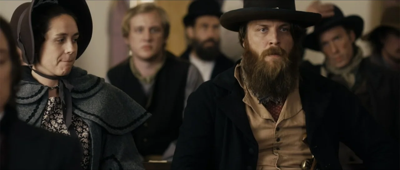 Out of Liberty (2019) movie still: medium — A group of people sit in a 19th-century meeting hall with wooden benches. A bearded man in a hat and a…