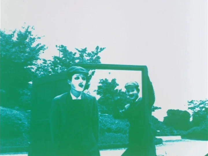 Labyrinth Tale (1975) movie still: medium — A man wearing white mime makeup and a flat cap stands in a park. He holds a large mirror that reflects…