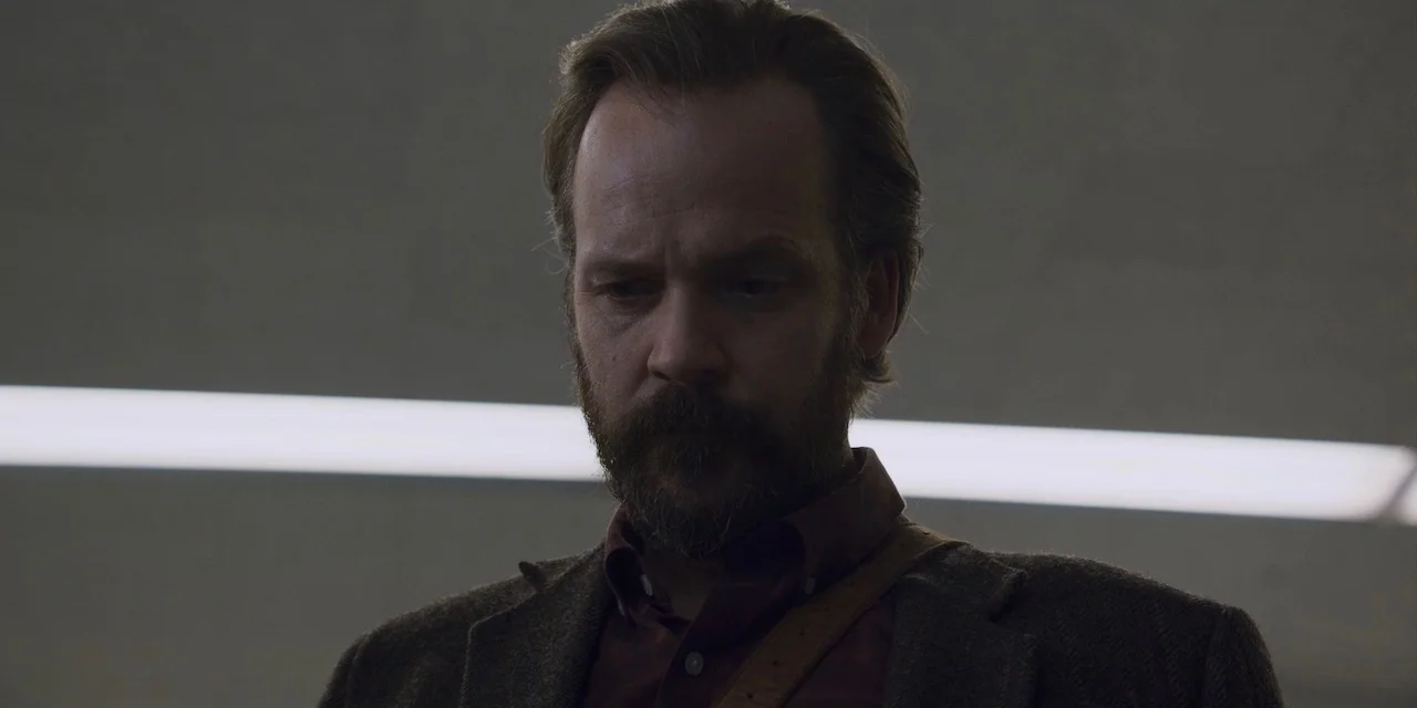The Sound of Silence movie still: closeup — A man stands in a sparsely lit interior space with a blank gray wall. The man gazes downward while…