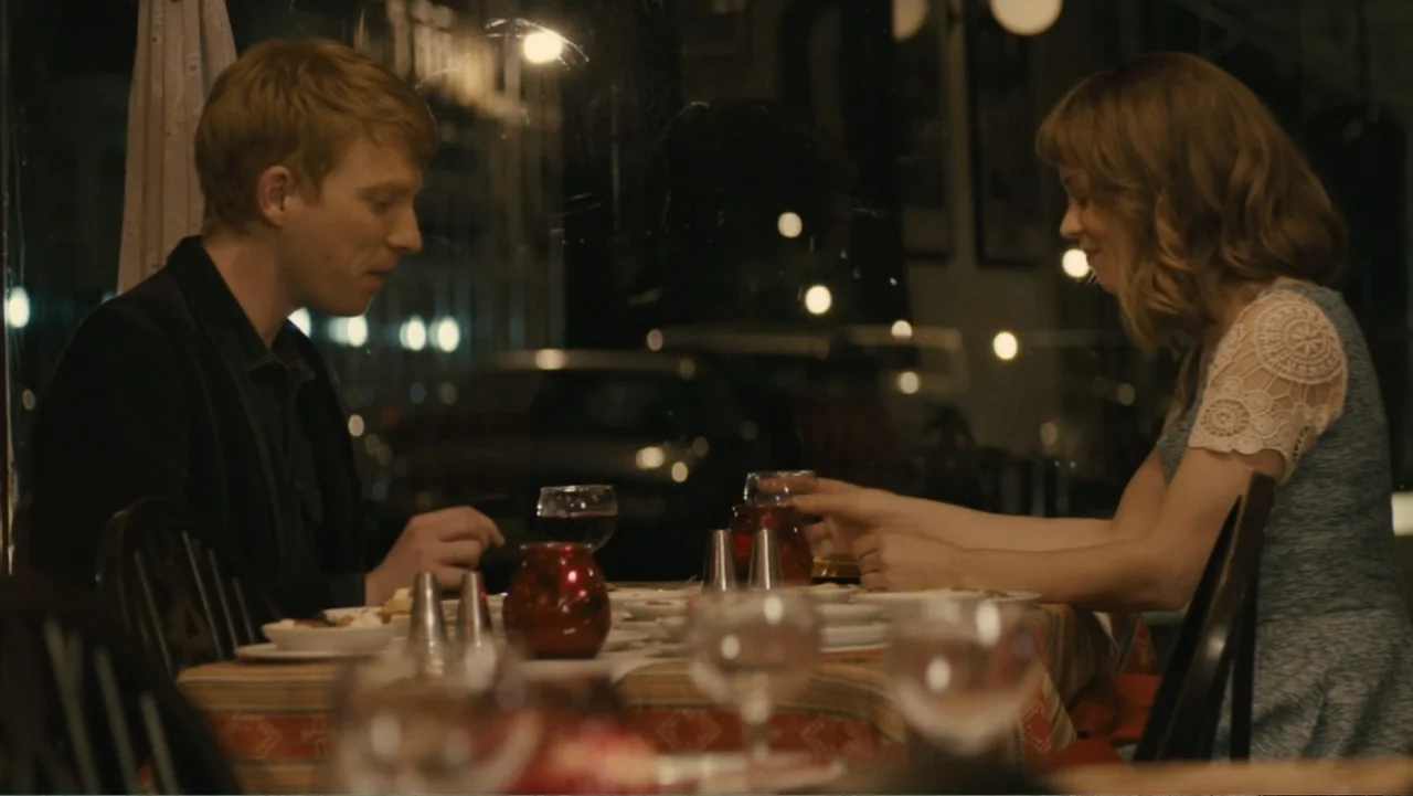 About Time (2013) movie still: medium — A dimly lit restaurant interior features a couple sitting at a table with dinnerware. The man and woman face…
