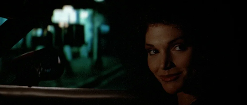 Scarface (1983) movie still: closeup — A dark car interior frames a woman looking out through the side window at night. Blurry city lights glow in…