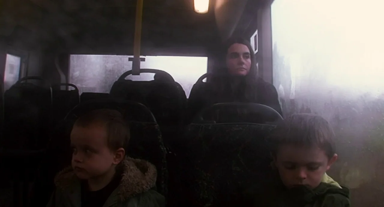 Everyday (2012) movie still: medium — The interior of a bus with patterned seats and rain-covered windows. A woman and two young boys sit quietly…