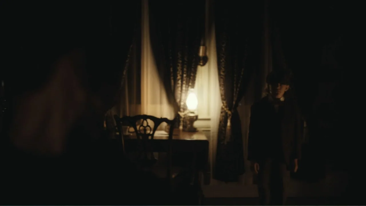 Demon in the White House (2021) movie still: medium — A dark, dimly lit room features a wooden chair and curtains near a window. A young boy wearing…