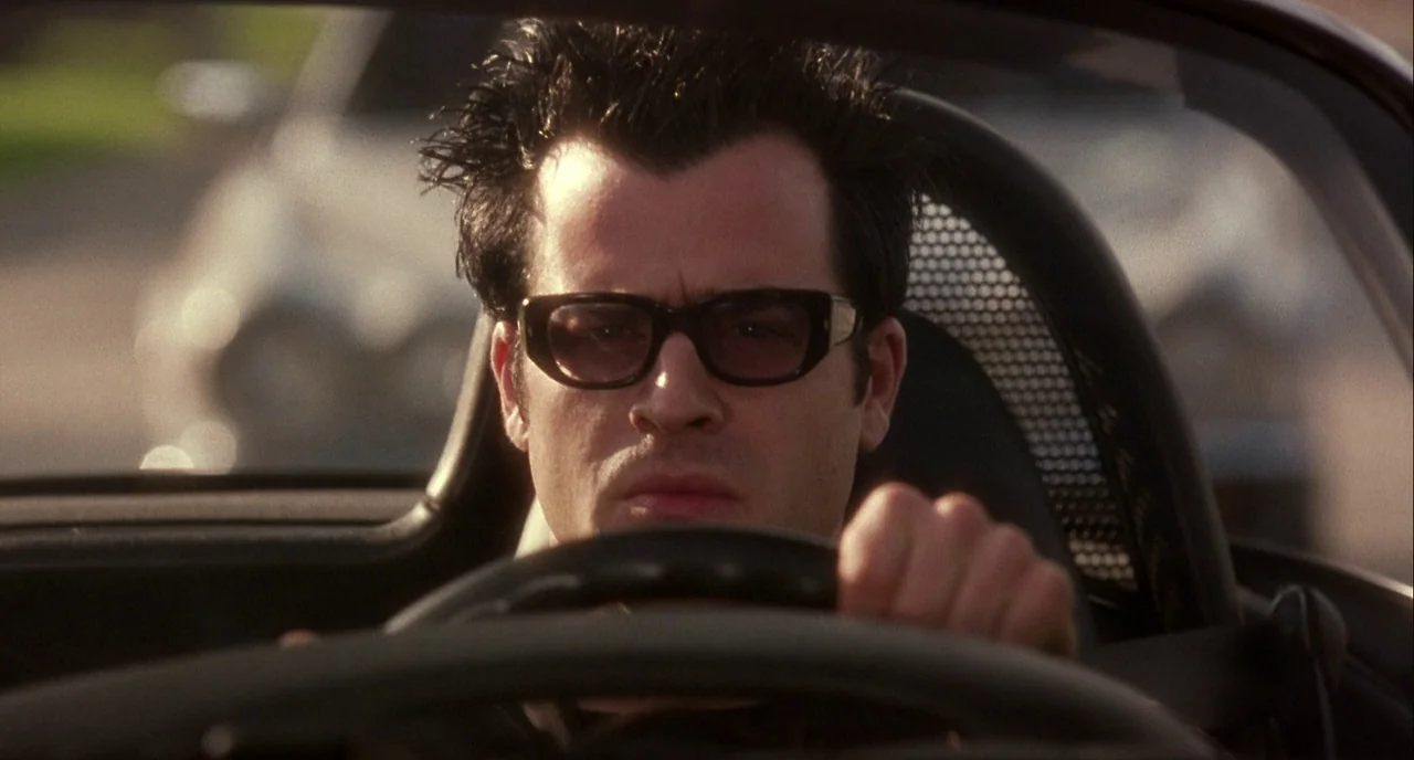 Mulholland Drive (2001) movie still: closeup — A man drives a convertible car along a road during the day. He grips the steering wheel while looking…