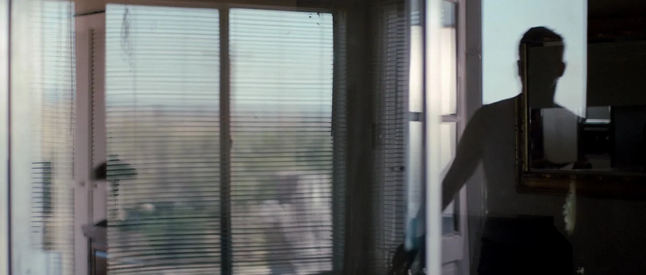Cop Car (2015) movie still: medium — A dimly lit room features a large window covered by horizontal plastic blinds. A shirtless man stands in…