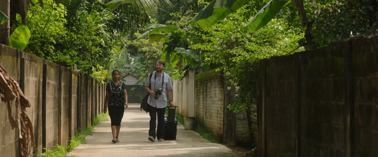 Greed (2019) movie still: fullbody — A narrow residential path bordered by concrete block walls and dense tropical vegetation. A woman and a man…