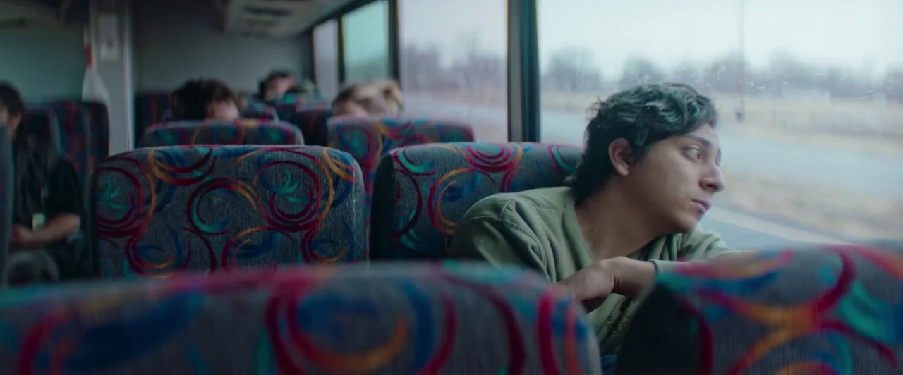 The Long Dumb Road (2018) movie still: medium — An interior of a bus with patterned seats showing several passengers in the background. A young man…