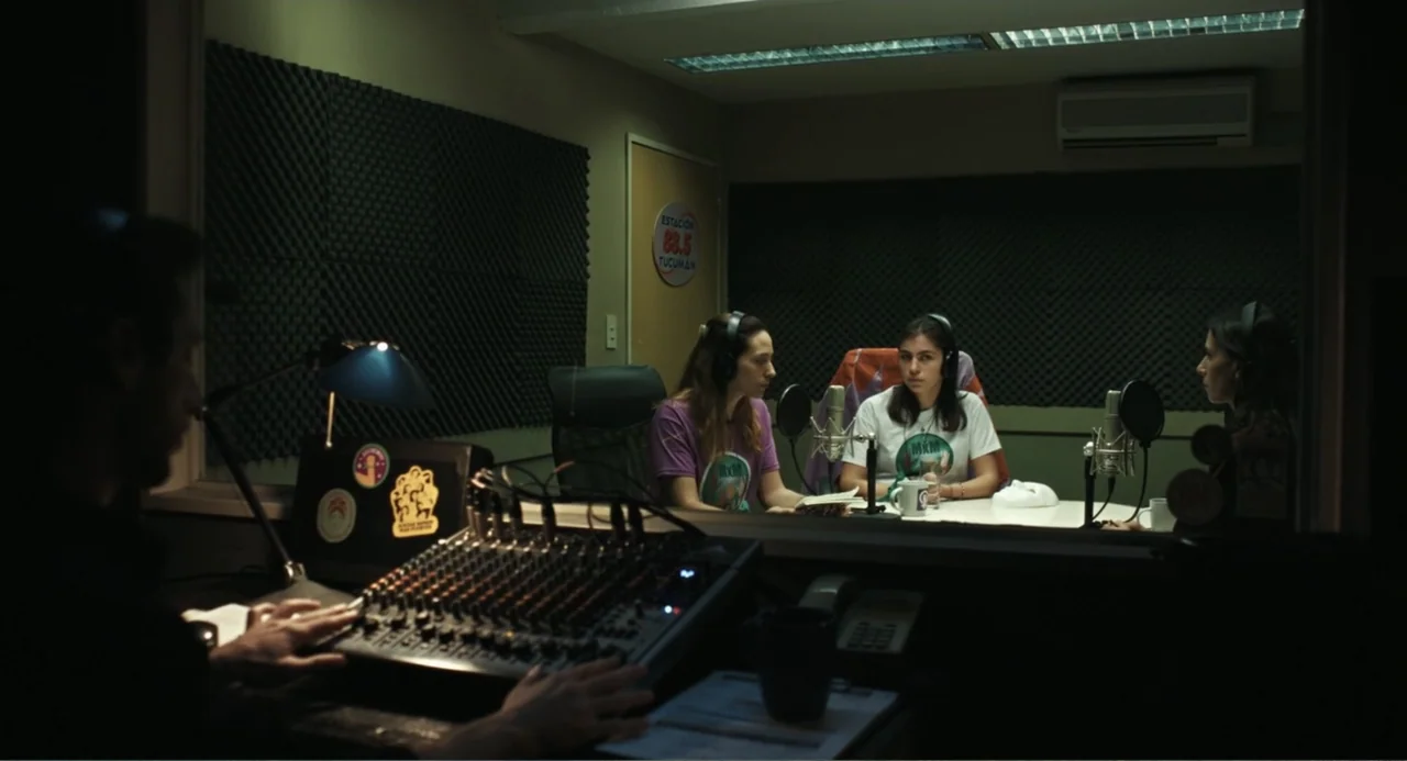 Belén (2025) movie still: medium — A radio studio booth viewed through a glass window with acoustic foam on walls. Two women sit at a desk with…