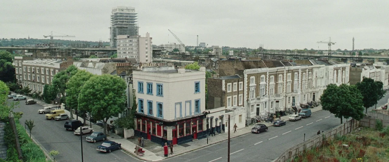 Mangrove (2020) movie still: establishing — An aerial view of a London residential street with terraced houses and a pub. Vintage cars are parked…