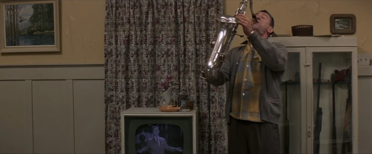 This Boy's Life (1993) movie still: medium — A living room features beige walls adorned with framed art and floral patterned curtains. A man stands…