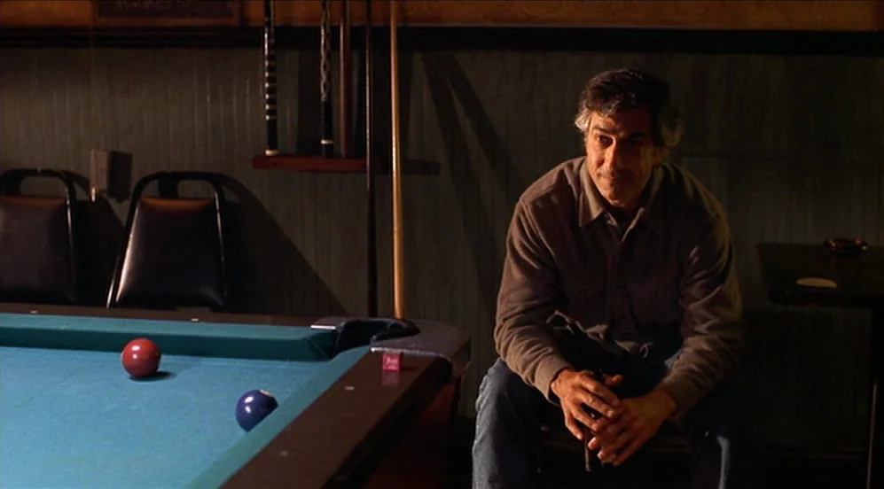 Limbo (1999) movie still: medium — A dimly lit pool hall with green felt tables and wooden wall paneling. An older man in a brown shirt sits quietly…