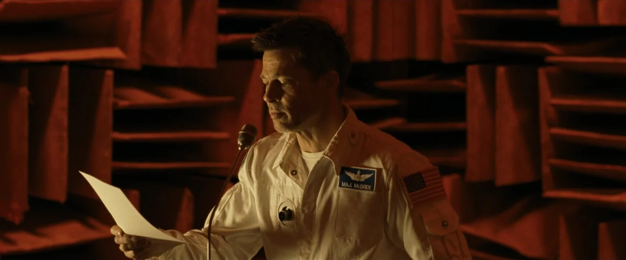 Ad Astra (2019) movie still: medium — A soundproof booth with rows of orange acoustic foam lining the rear walls. A man in a flight suit holds a…