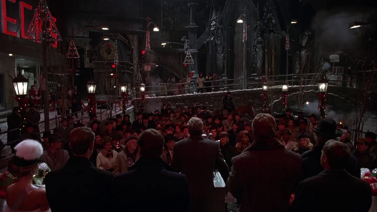 Batman Returns (1992) movie still: wide — A dense crowd gathers in a dark, Gothic-inspired town square during a winter evening. Many people face…