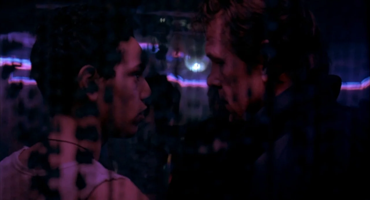 The Good Thief (2002) movie still: medium — Two men stand face-to-face inside a dark club lit by magenta neon lighting. The camera is partially…
