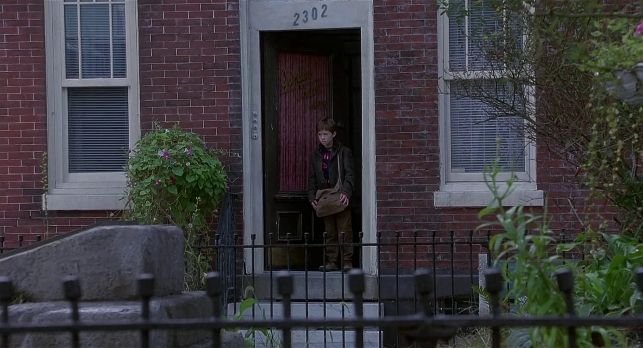 The Sixth Sense (1999) movie still: wide — A red brick row house features a black door labeled 2302. A young boy stands inside the doorway.