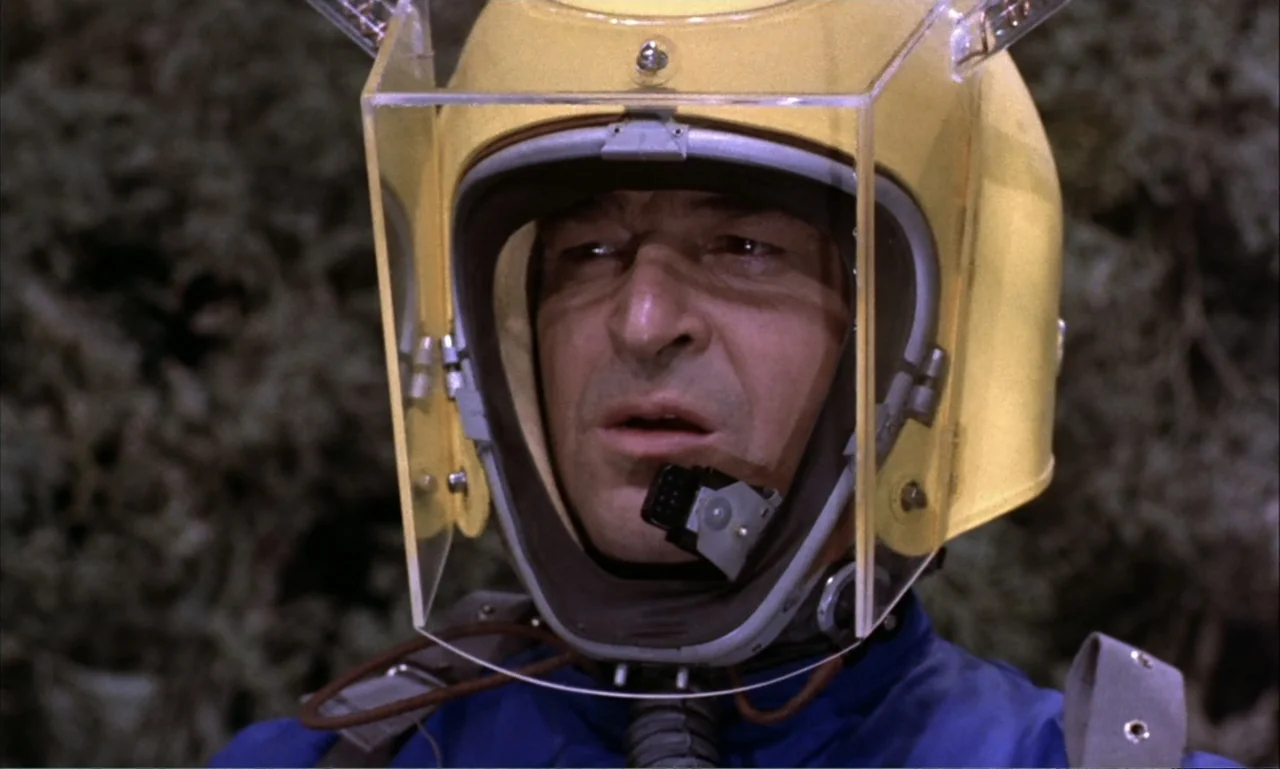 Journey to the Seventh Planet (1962) movie still: closeup — An outdoor location with blurry green foliage in the background. A man wears a yellow…