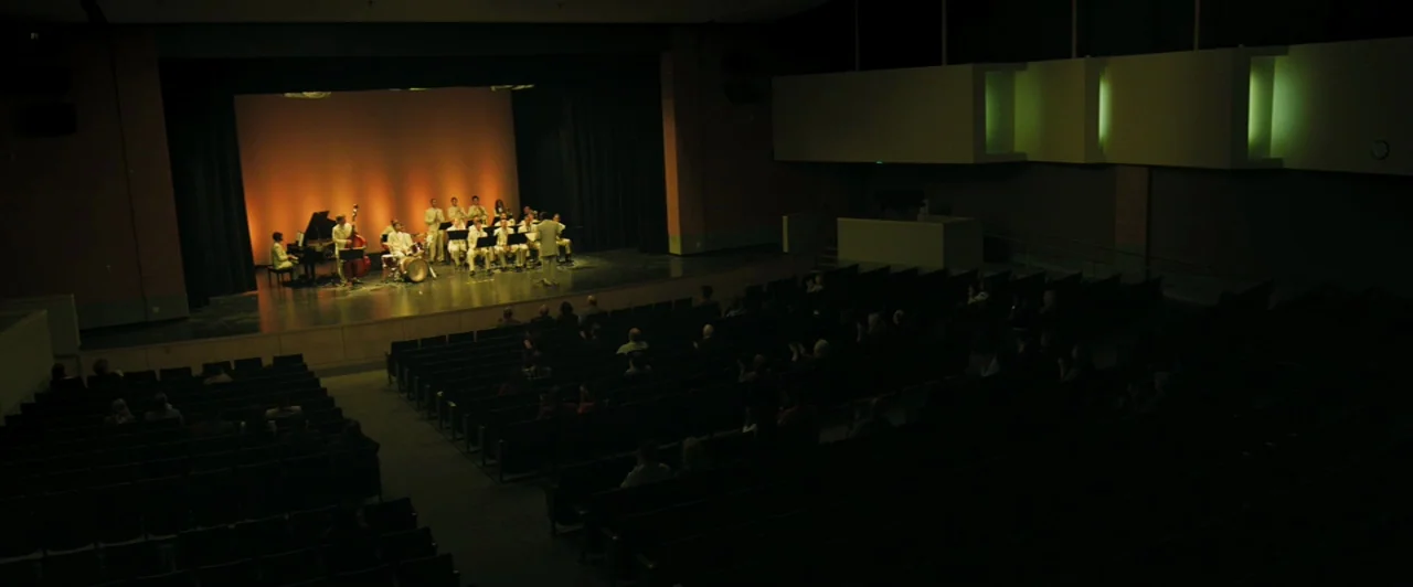 Whiplash (2014) movie still: wide — A large dark auditorium features rows of empty seats facing an illuminated wooden stage. A band dressed in white…