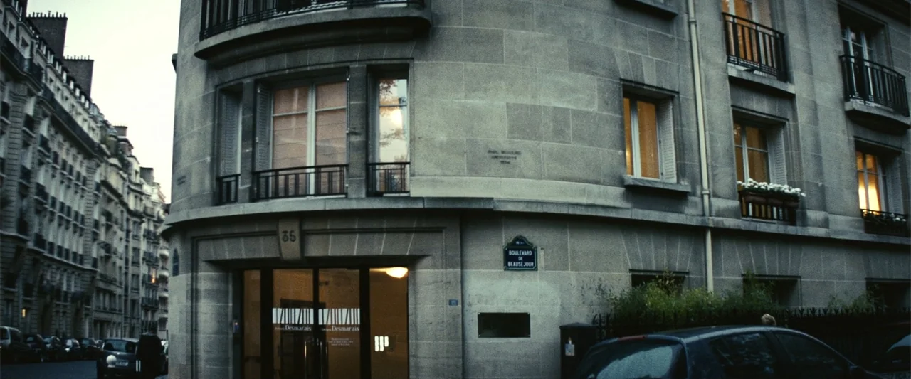 Hereafter (2010) movie still: wide — A curved stone apartment building stands on a street corner during the twilight hour. Dark windows and lit…