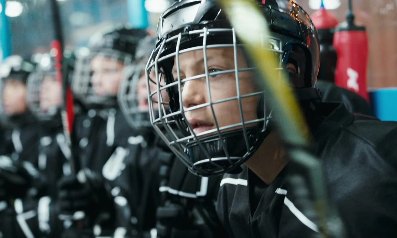 Close (2022) movie still: closeup — A row of youth hockey players sits on a team bench behind a plexiglass barrier. The lead boy in the foreground…