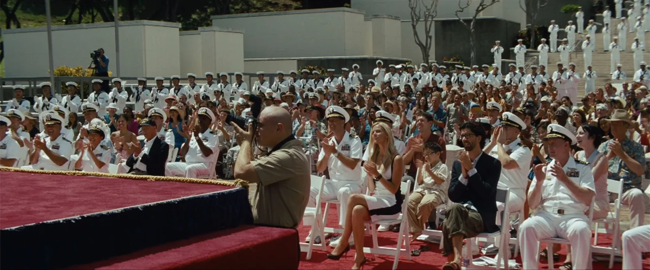 Battleship (2012) movie still: wide — A large outdoor audience sits on white chairs facing a red carpeted stage. Many individuals in United States…