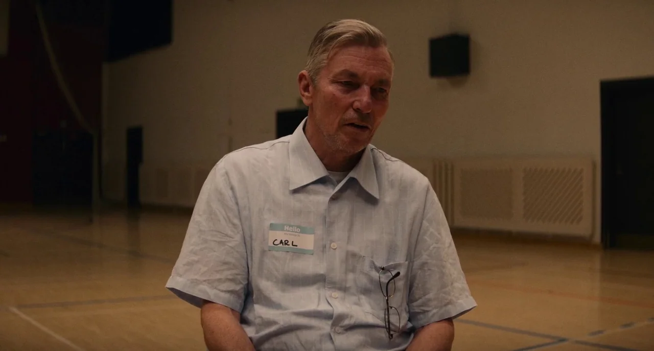 Dual (2022) movie still: medium — A gymnasium interior features wooden floors and muted walls under dim, soft lighting. An older man wearing a name…