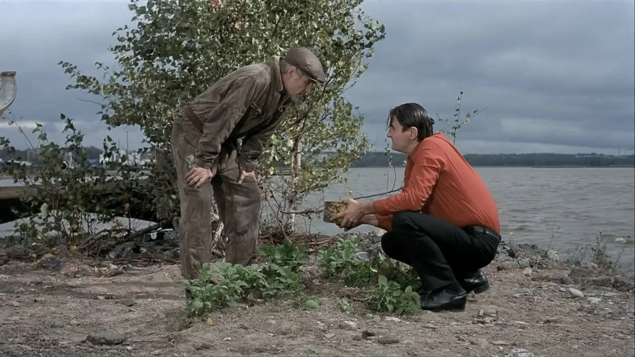 The Man Without a Past (2002) movie still: fullbody — A rocky, sparsely vegetated lakeshore sits under a gray, overcast sky. Two men crouch near the…