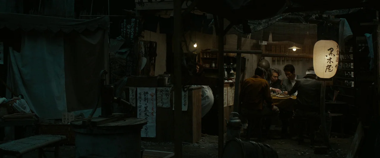 Godzilla Minus One (2023) movie still: wide — A dimly lit outdoor stall features handwritten paper signs and a glowing paper lantern. Three men sit…