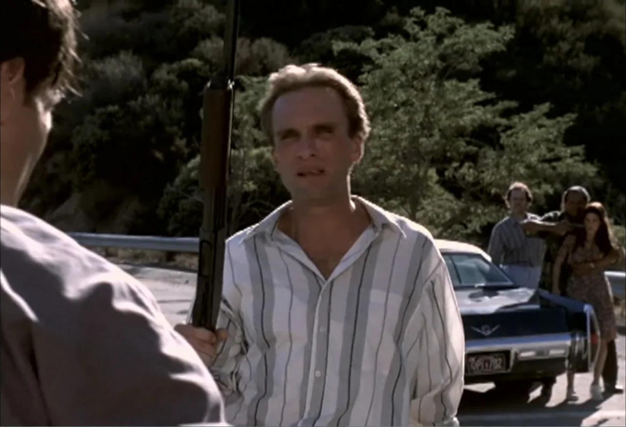 Black Cat Run (1998) movie still: medium — A rural mountain road features a parked vintage sedan near a steep hillside. A man in a striped shirt…