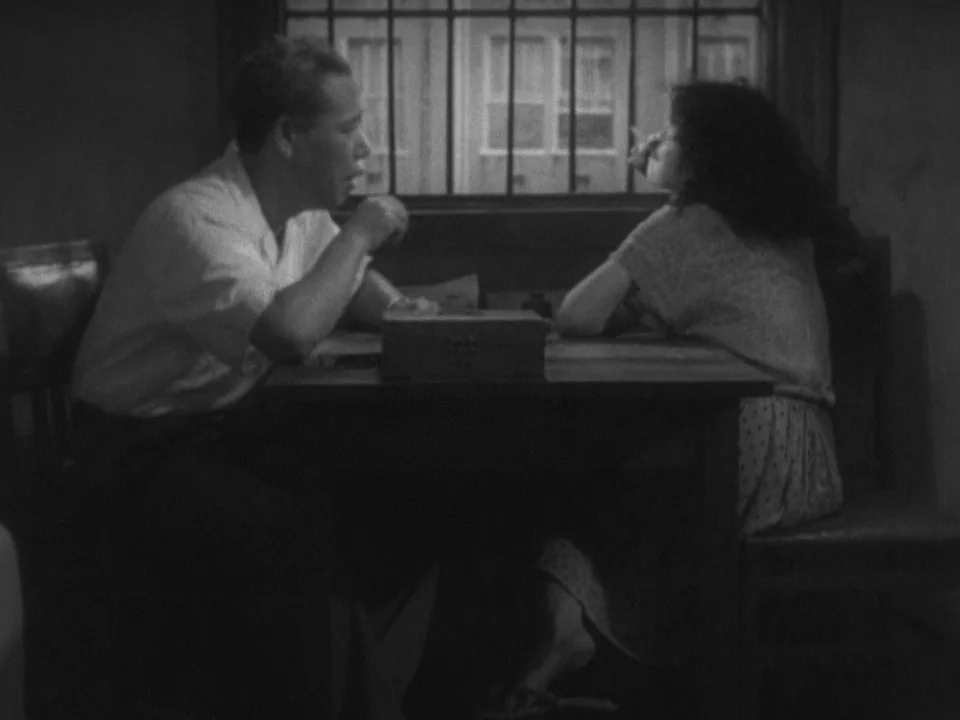 Stray Dog (1949) movie still: medium — A dark, dimly lit room contains a table with two people sitting at it. A window with iron bars and curtains…