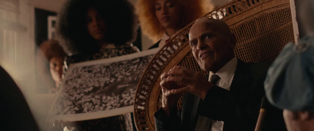 BlacKkKlansman (2018) movie still: medium — A group of people gathers in a residential room filled with textured furniture and art. An elderly man…