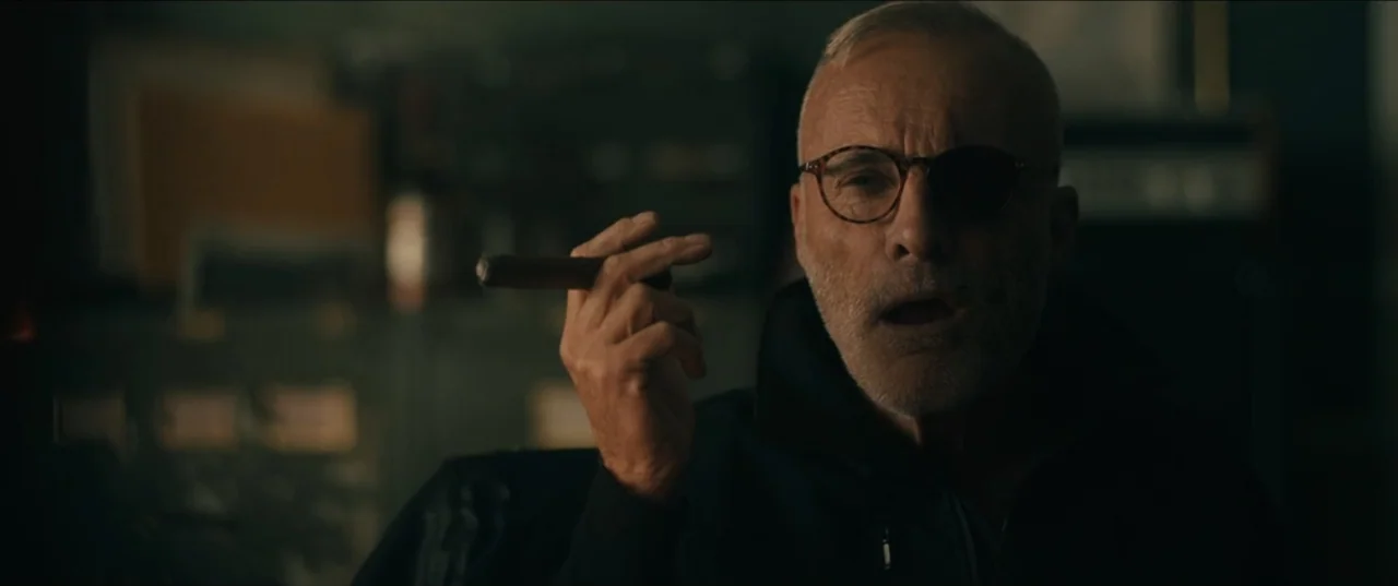 Guns Up (2025) movie still: closeup — A dark, dimly lit office interior contains indistinct furniture and shelves in the background. An older man…