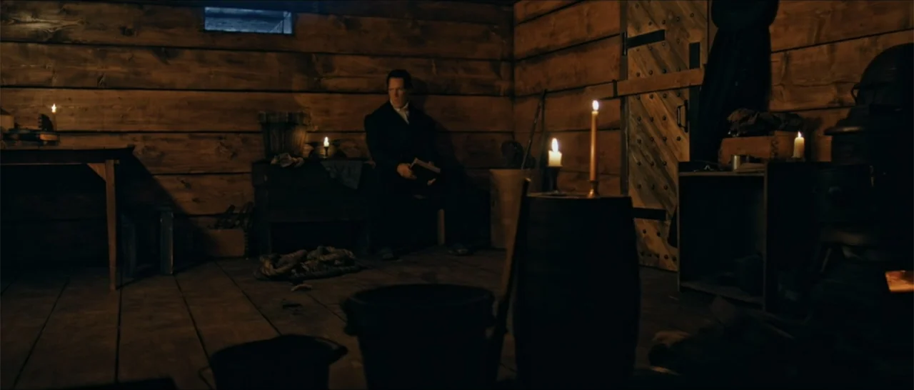 Out of Liberty (2019) movie still: medium — A rustic interior with log walls and wooden floors dimly lit by scattered candles. A solitary man in…