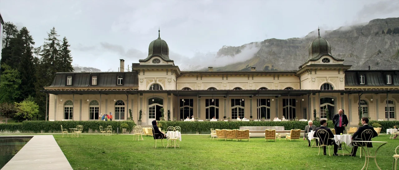 Youth (2015) movie still: establishing — A historic, grand hotel building with multiple domes sits against a backdrop of mountains. People are…